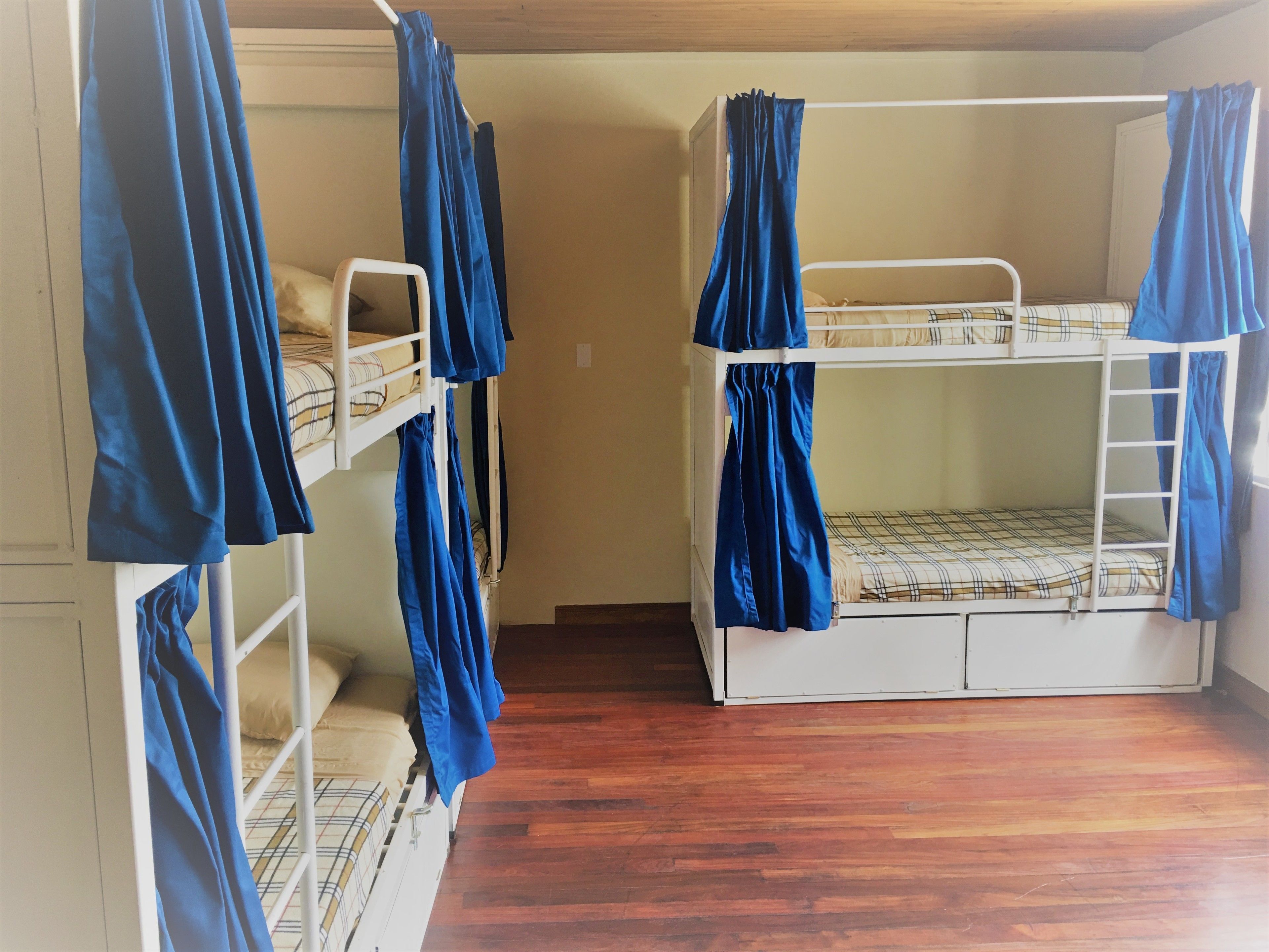 Ein Zimmer mit zwei Etagenbetten, jeweils ausgestattet mit einer Matratze und einer Decke. Der Boden ist aus Holz und die Wand weiß.
