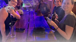 Eine Gruppe von Personen sitzt an einem Tisch in einer Bar mit tropischem Wandbild im Hintergrund, spielt Karten und hält Getränke.