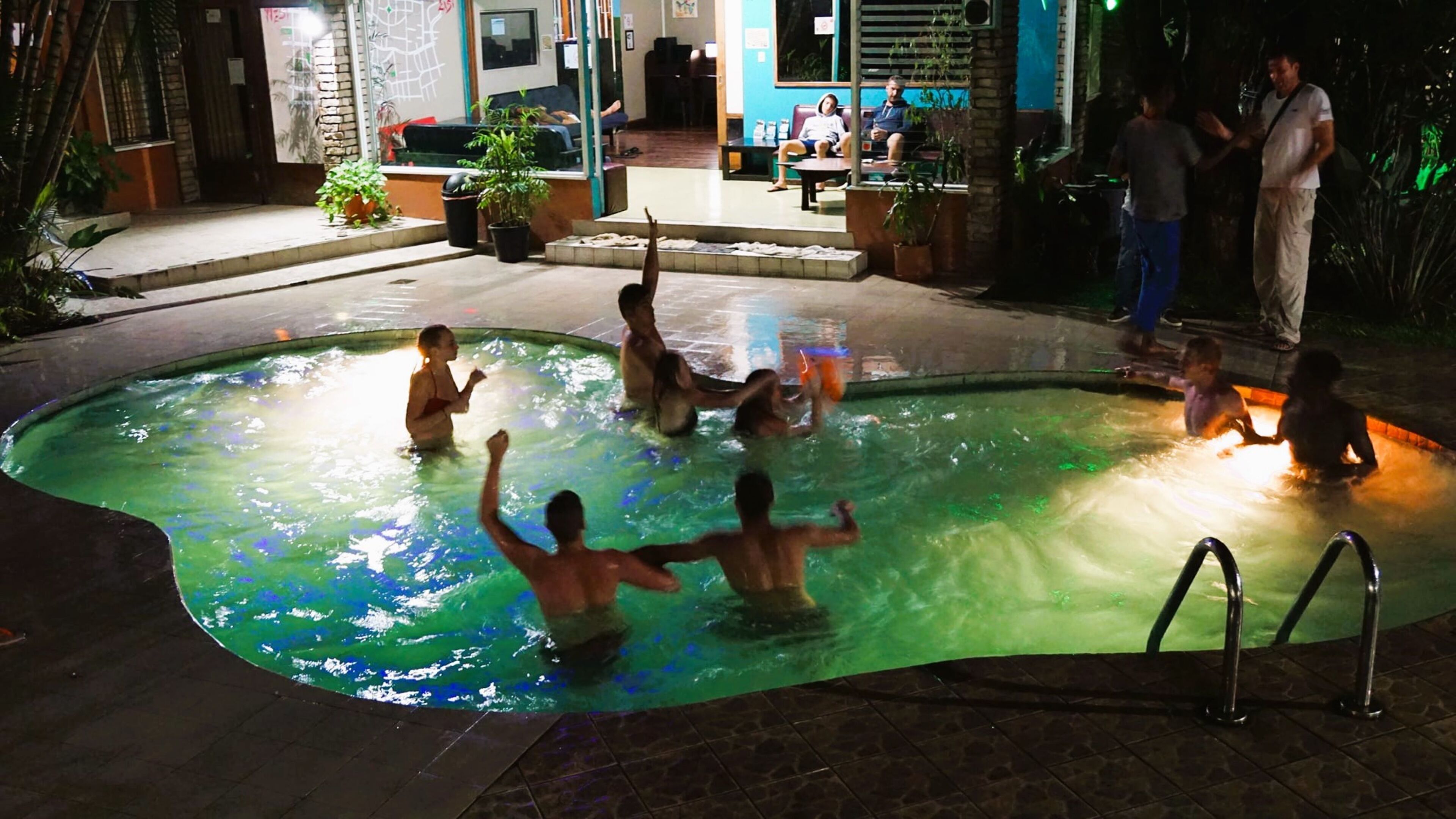 A swimming pool area with a tiled deck, a few potted plants, and a seating area with chairs and tables.
