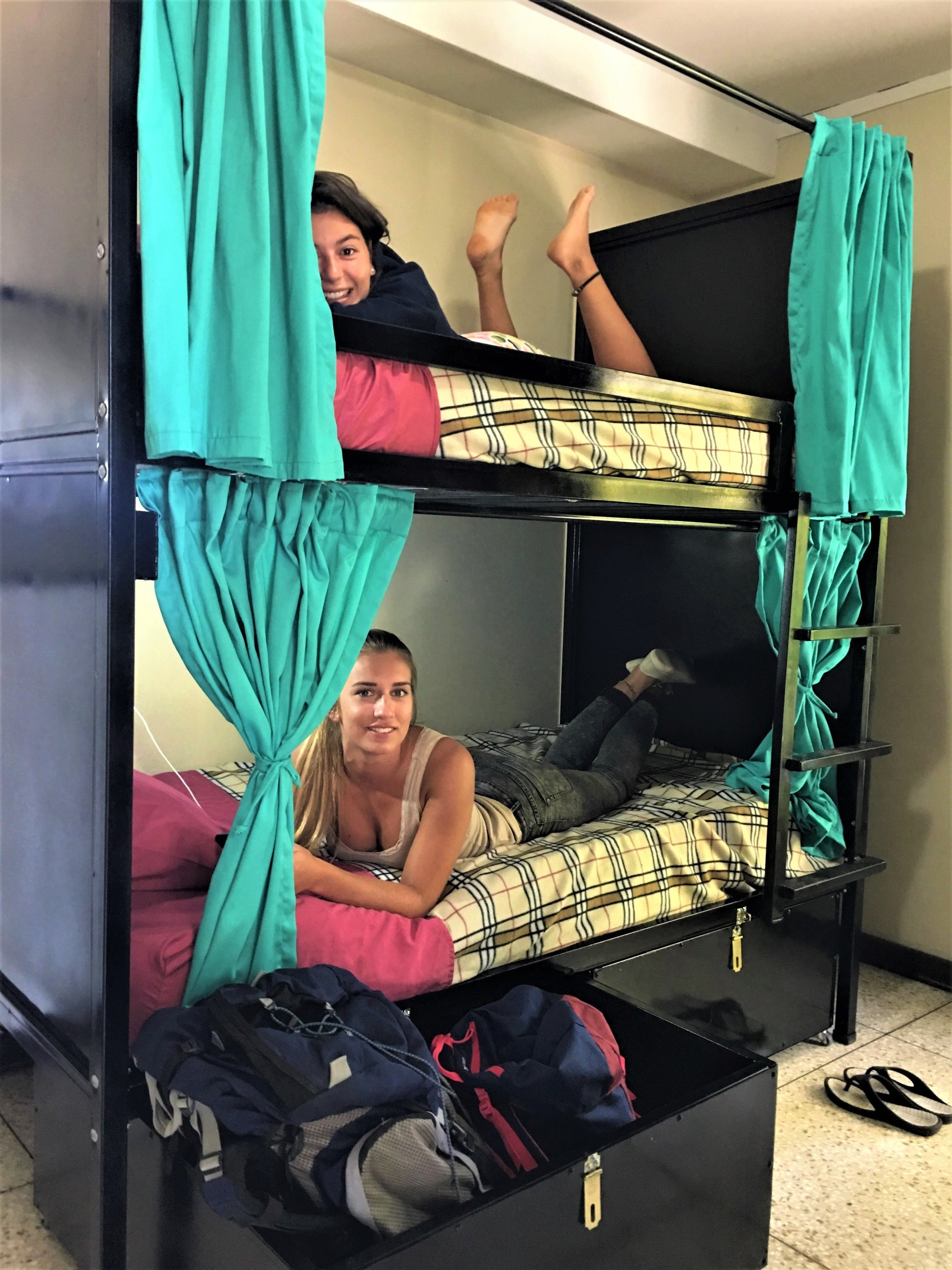 Una cama litera con cortinas turquesa, una cama con ropa de cama escocesa y una maleta en el suelo.