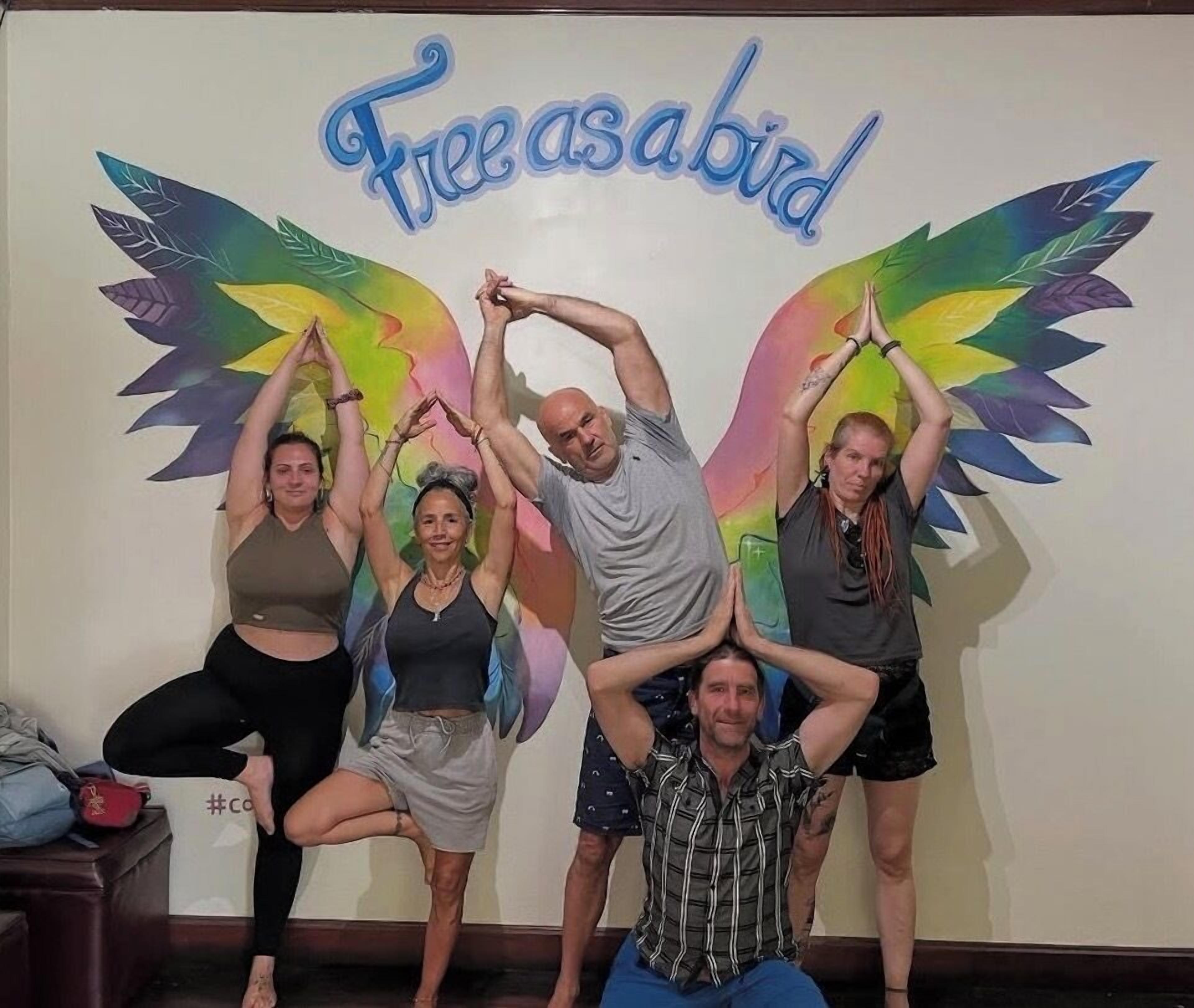 Un groupe de personnes effectuant une posture de yoga devant un mur orné d’un motif d’oiseau coloré et sur lequel est inscrit le texte : « Libre comme l’air ».