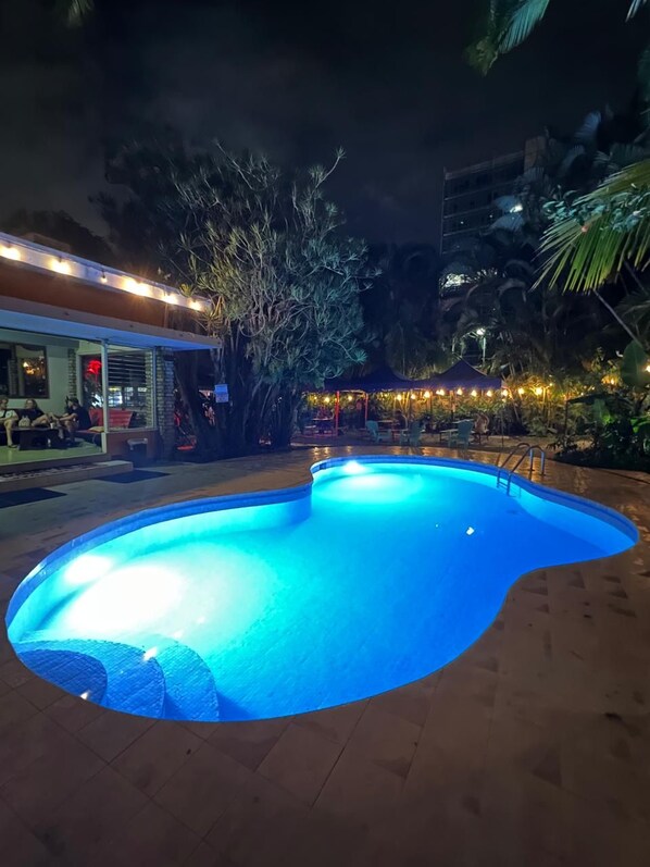 A swimming pool with a curved edge, surrounded by a paved area. There is a building with a glass facade and outdoor seating visible in the background.