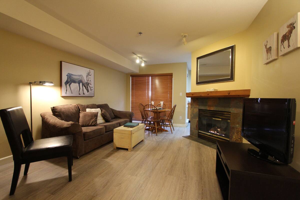 Una sala de estar con un sofá marrón, una mesa de centro de madera, un televisor y una chimenea.