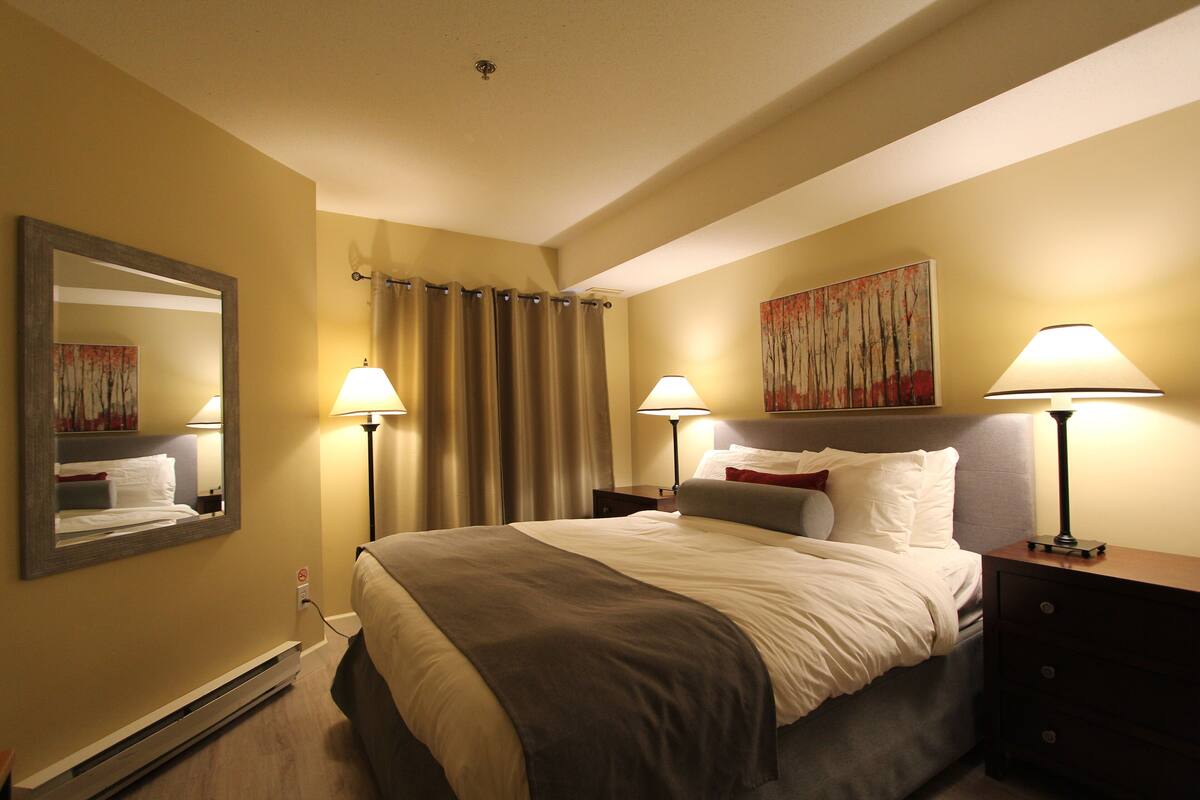 Una habitación de hotel con cama, dos mesitas de noche, un espejo, un cuadro y un calentador.