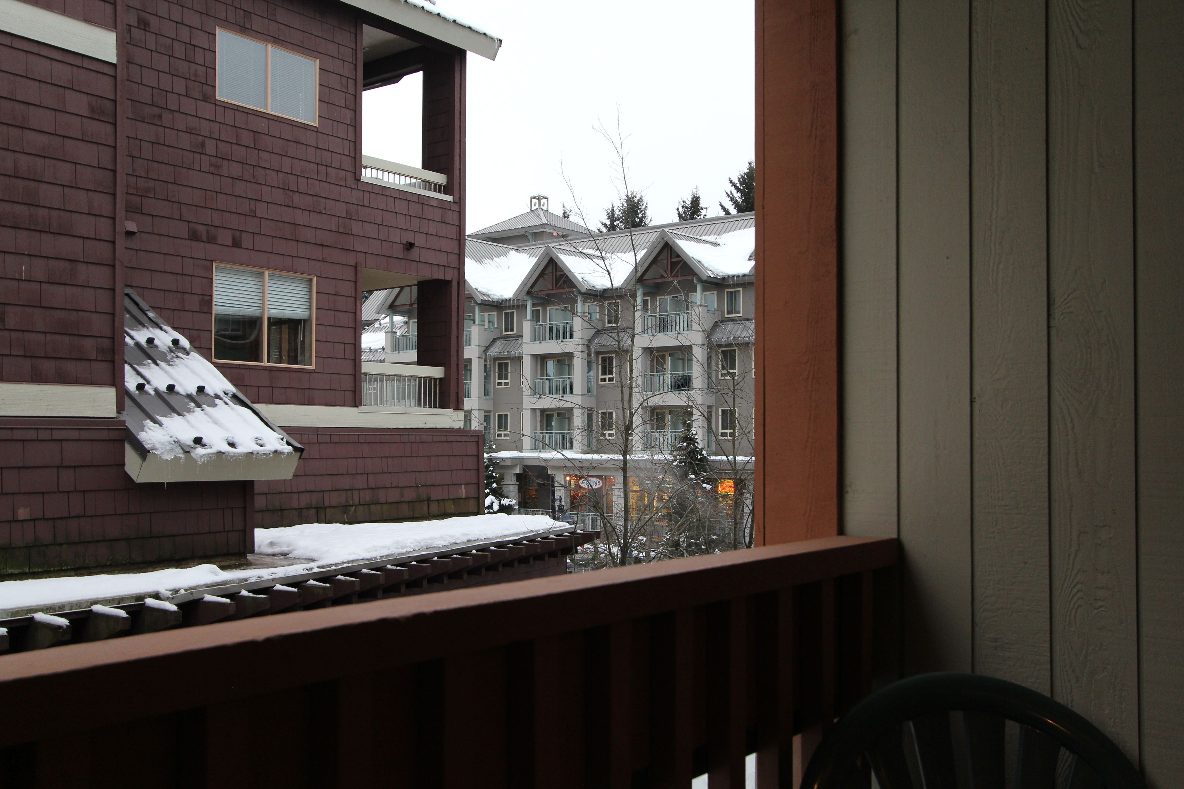 A view from a balcony with a snow-covered roof and a wooden railing. In the background, there are multi-story buildings with balconies and a snowy landscape.