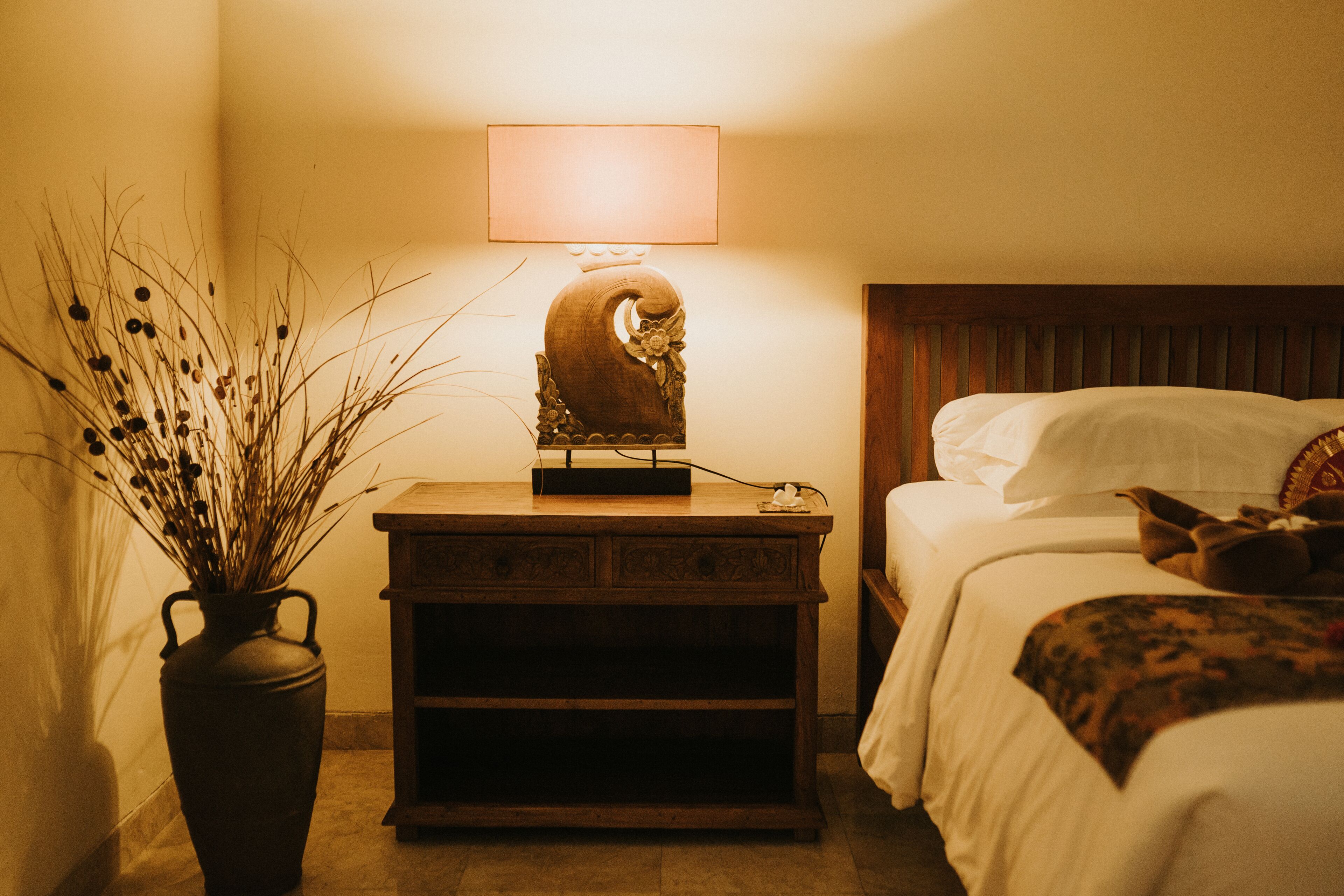 Ein Schlafzimmer mit Nachttisch aus Holz, einer Vase mit Trockenpflanzen und einem Bett mit weißen Leinentüchern und einer gemusterten Tagesdecke.