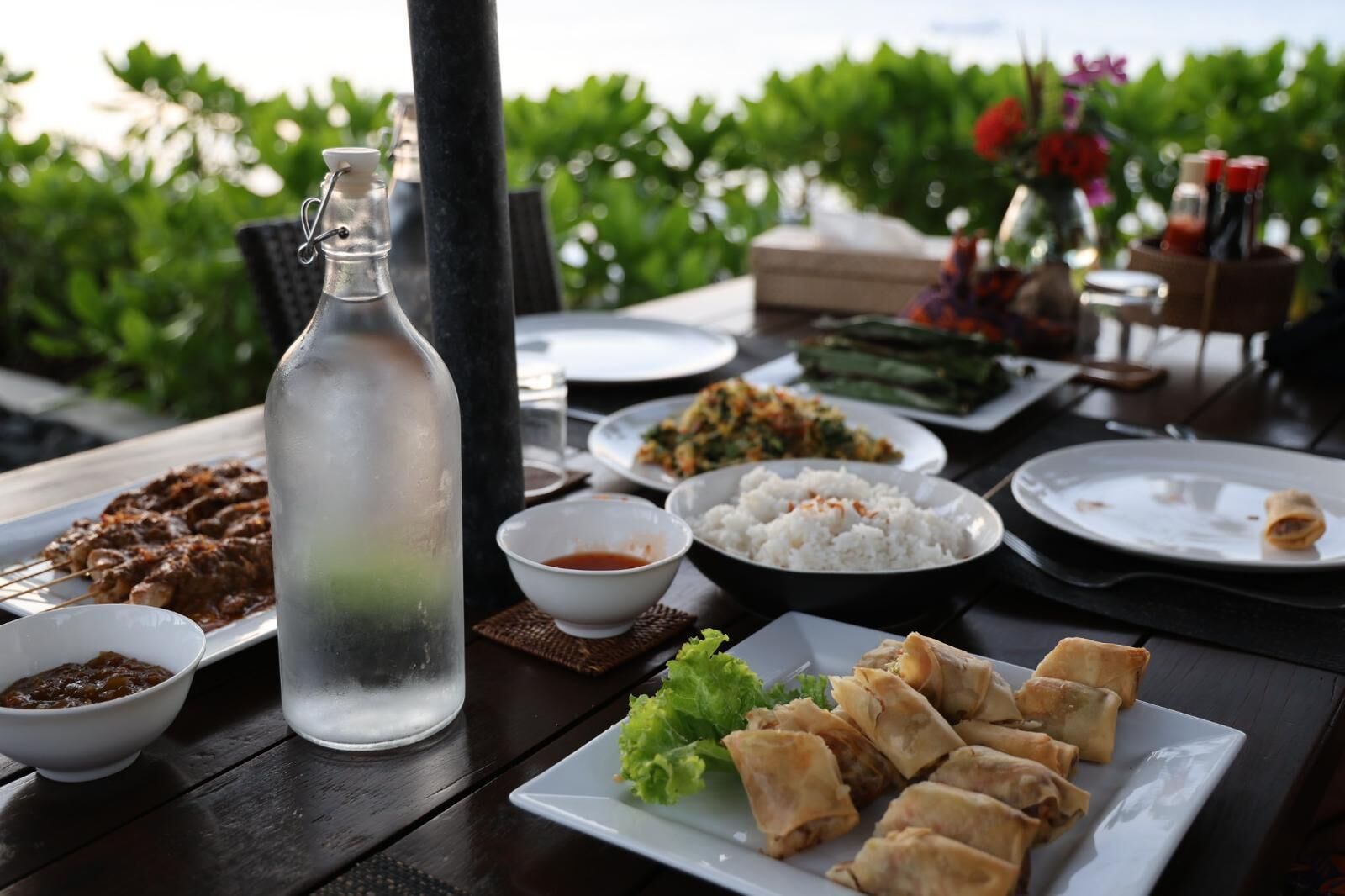 A table set with various dishes including rice, meat, and spring rolls, with a bottle of water and condiments.
