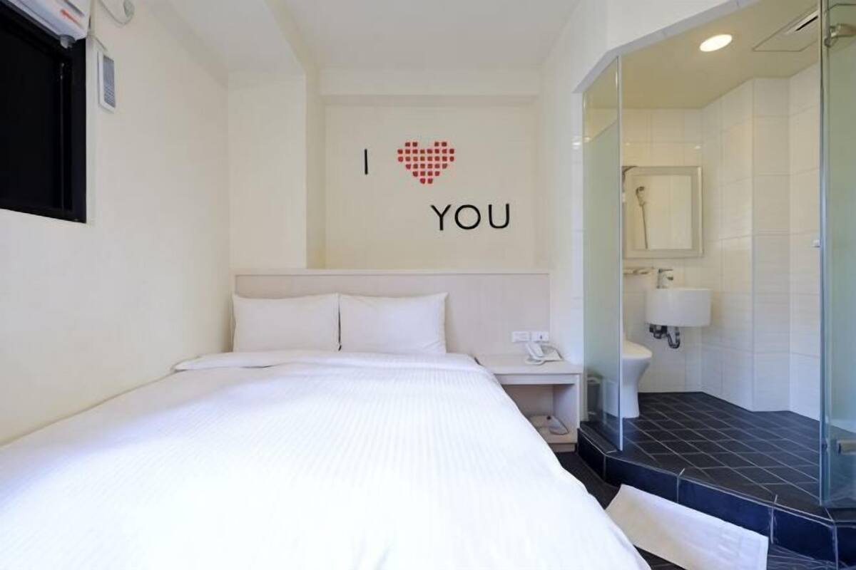 A hotel room with a bed, a bedside table, a phone, a mirror, and a shower area.