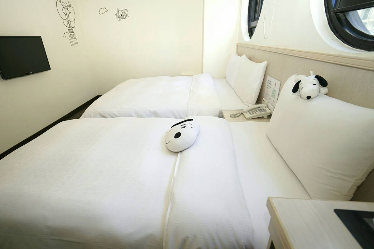 A hotel room with two beds, a wall-mounted TV, and a window with a view of the sky.