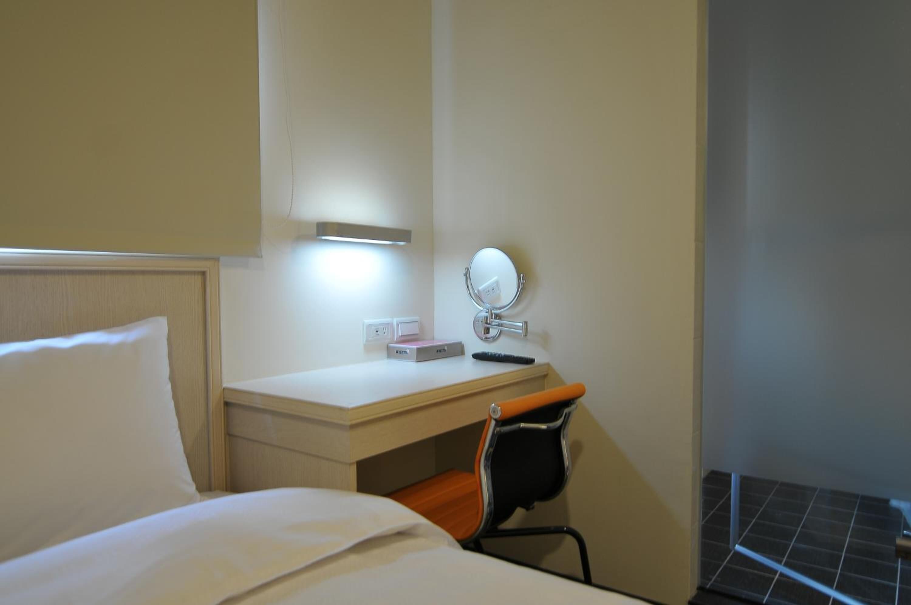 Een hotelkamer met een bed, een bureau met stoel, een klein tafeltje, een spiegel aan de muur en een lampje aan de muur.