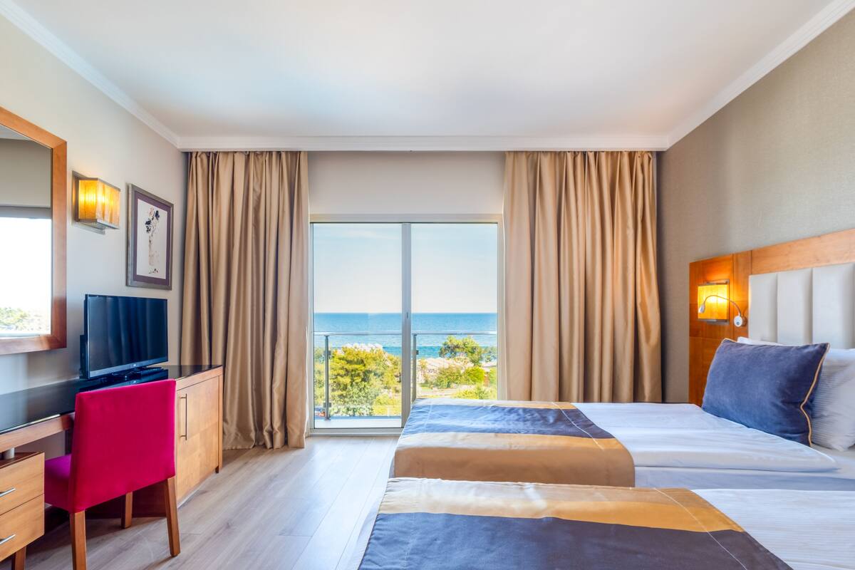 A hotel room with a bed, a desk with a computer, a chair, and a large window with curtains.