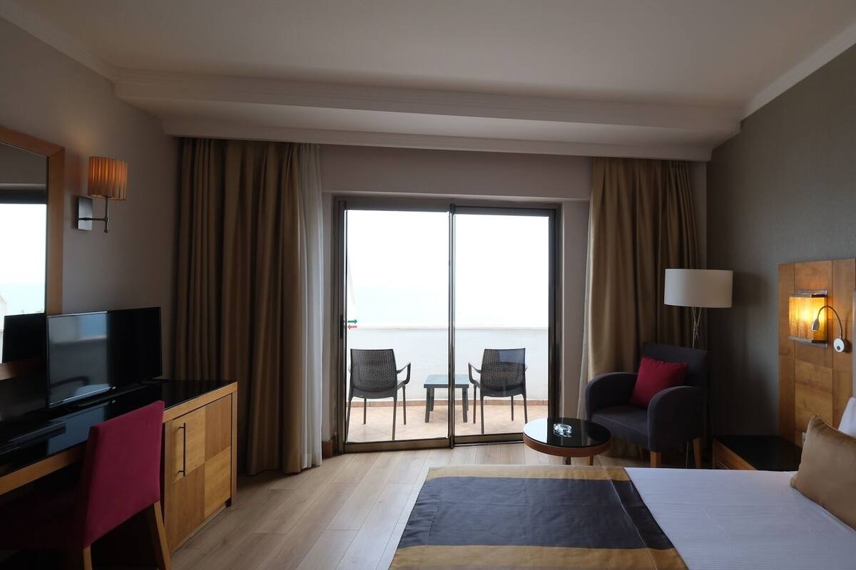 A hotel room with a bed, a desk with a TV, a chair, a small table, and a view of the sea through the sliding doors.