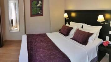 A neatly made bed with a white and purple bedspread, two pillows, and a headboard with wall-mounted lamps.