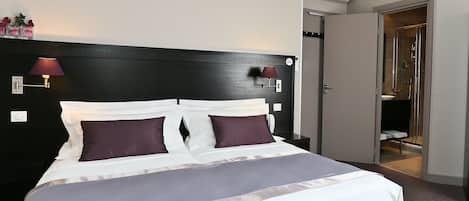 Una cama bien hecha con ropa de cama blanca y morada, un cabecero negro y dos lámparas fijadas a la pared.