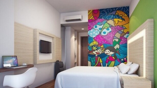 Een moderne hotelkamer met een bed, een bureau met een laptop, een televisie en een kleurrijke muurschildering.