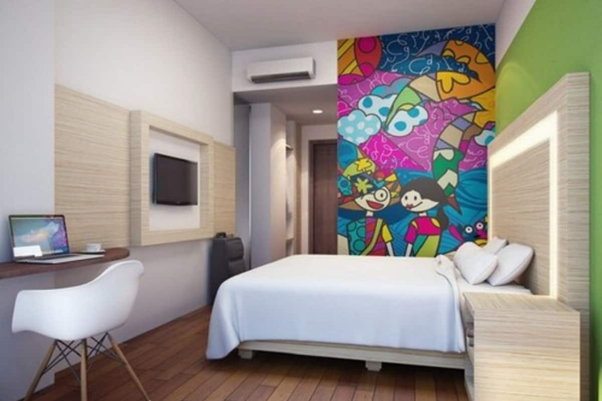 Een moderne hotelkamer met een bed, een bureau met een laptop, een televisie en een kleurrijke muurschildering.