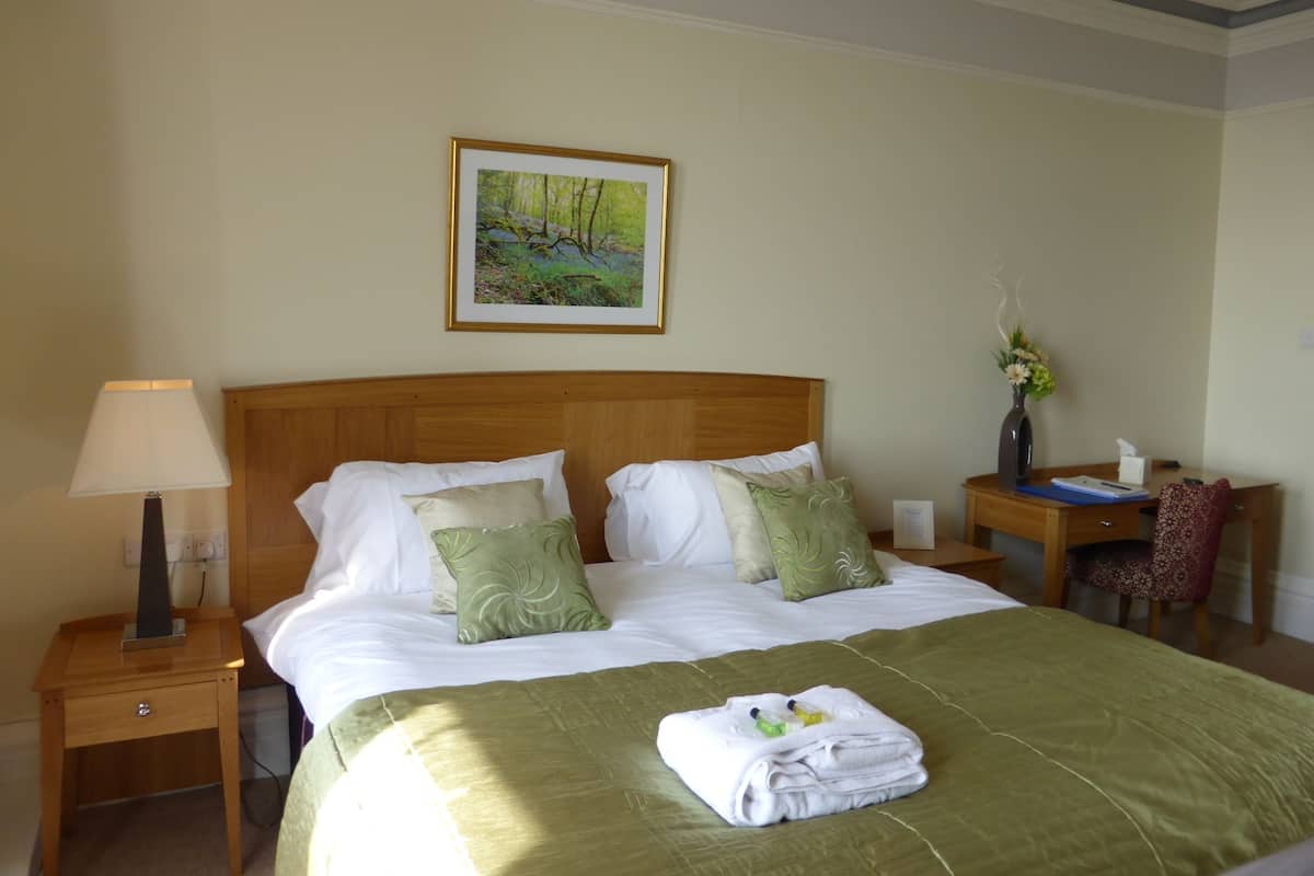 A neatly made bed with a green bedspread, white linens, and decorative pillows. There is a wooden nightstand with a lamp and a small table with a vase of flowers. A framed picture hangs on the wall above the bed.