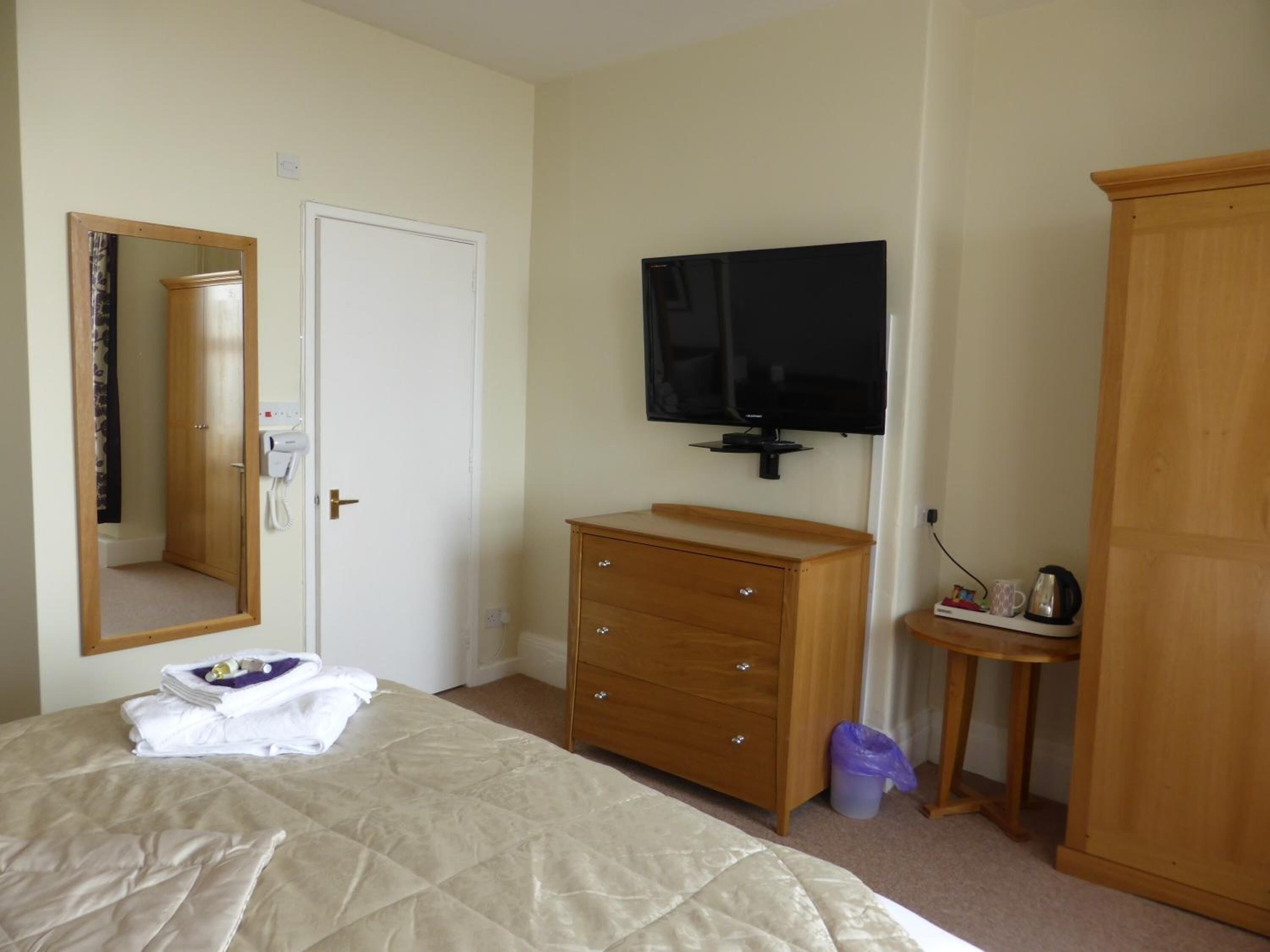 A hotel room with a bed, a wooden dresser, a television mounted on the wall, and a wardrobe.