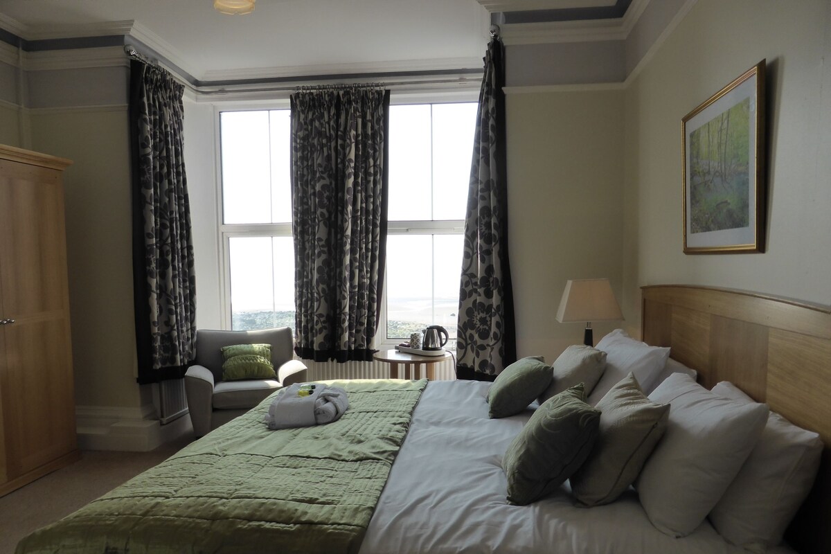 A hotel room with a large bed, a chair, a small table, and a window with curtains.