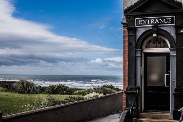 An entrance with a sign, a view of the ocean, and a building with a black door. - Bailey Ground Lodge (Seascale)