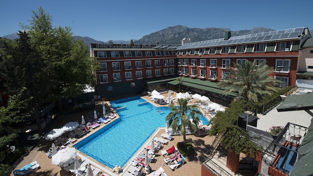 A hotel with a swimming pool surrounded by sun loungers and umbrellas, a palm tree, and mountains in the background. - Hotel Asdem Park (Kemer)