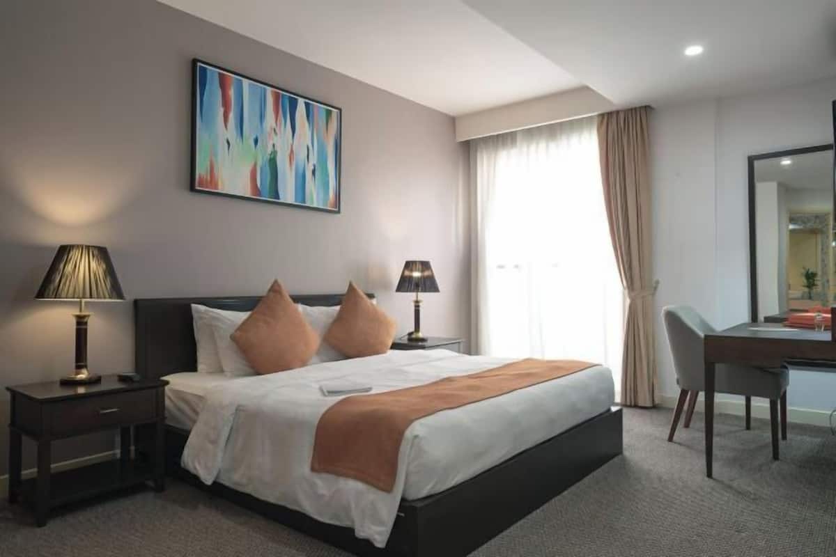Habitación de hotel con cama, dos mesitas de noche con lámparas, un escritorio con silla, un cuadro abstracto grande en la pared y una ventana con cortinas.