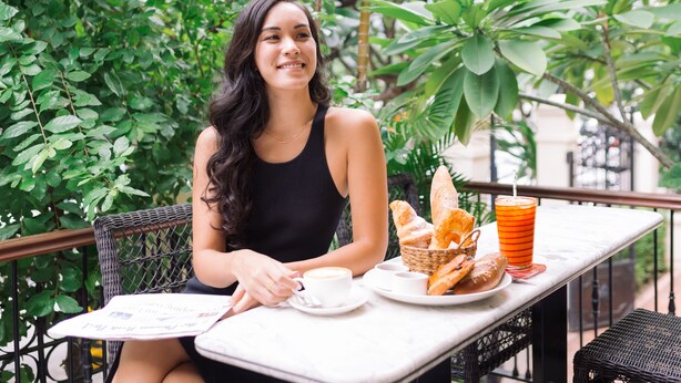 Una persona sentada en una mesa al aire libre, con una cesta de pan, una taza de café y un vaso de zumo de naranja.