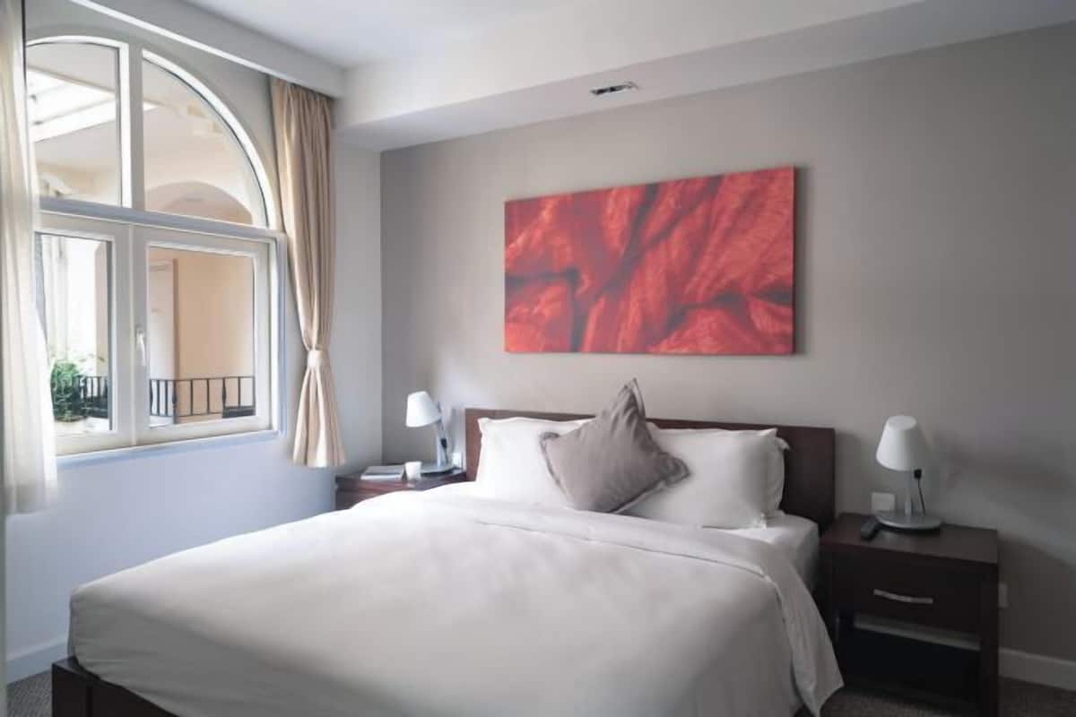 Una habitación de hotel con una cama grande, una mesita de noche con lámpara, una ventana con cortinas y un cuadro abstracto rojo en la pared.
