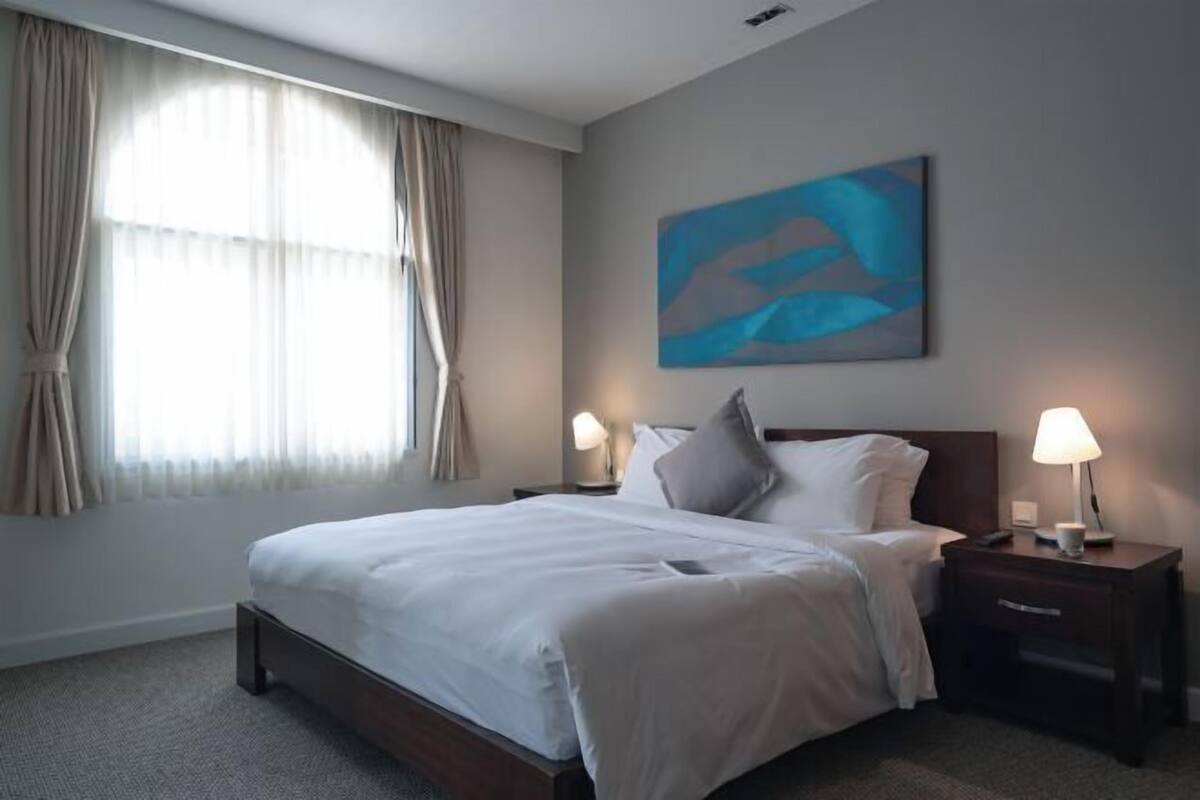 Una habitación de hotel con una cama, dos mesitas de noche con lámparas, un ventanal con cortinas y un cuadro abstracto colgado en la pared.