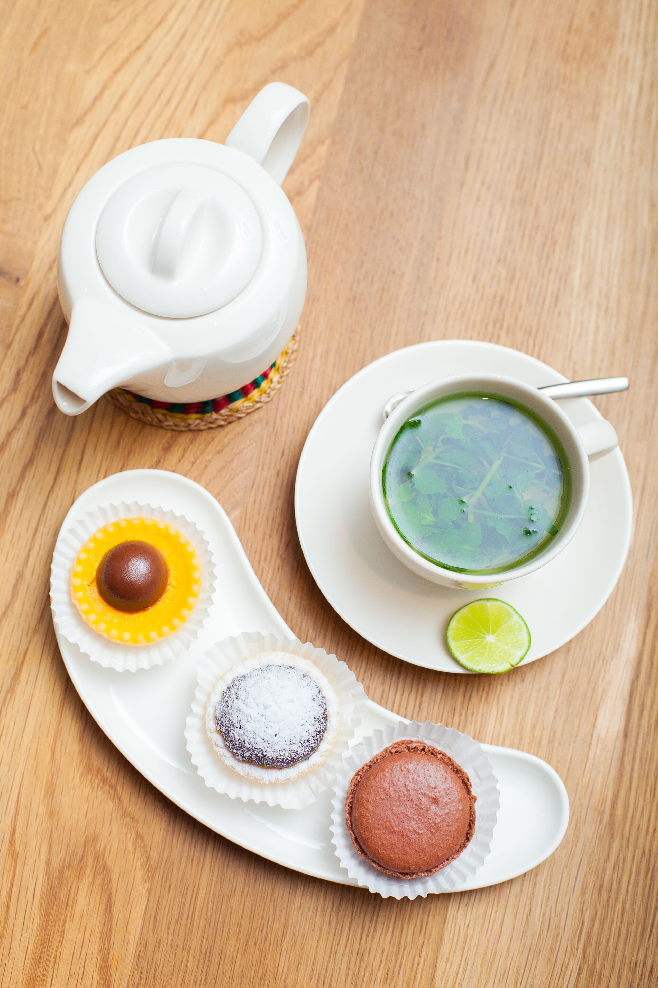 Una tetera blanca, un plato de sopa verde y tres pasteles sobre una mesa de madera.