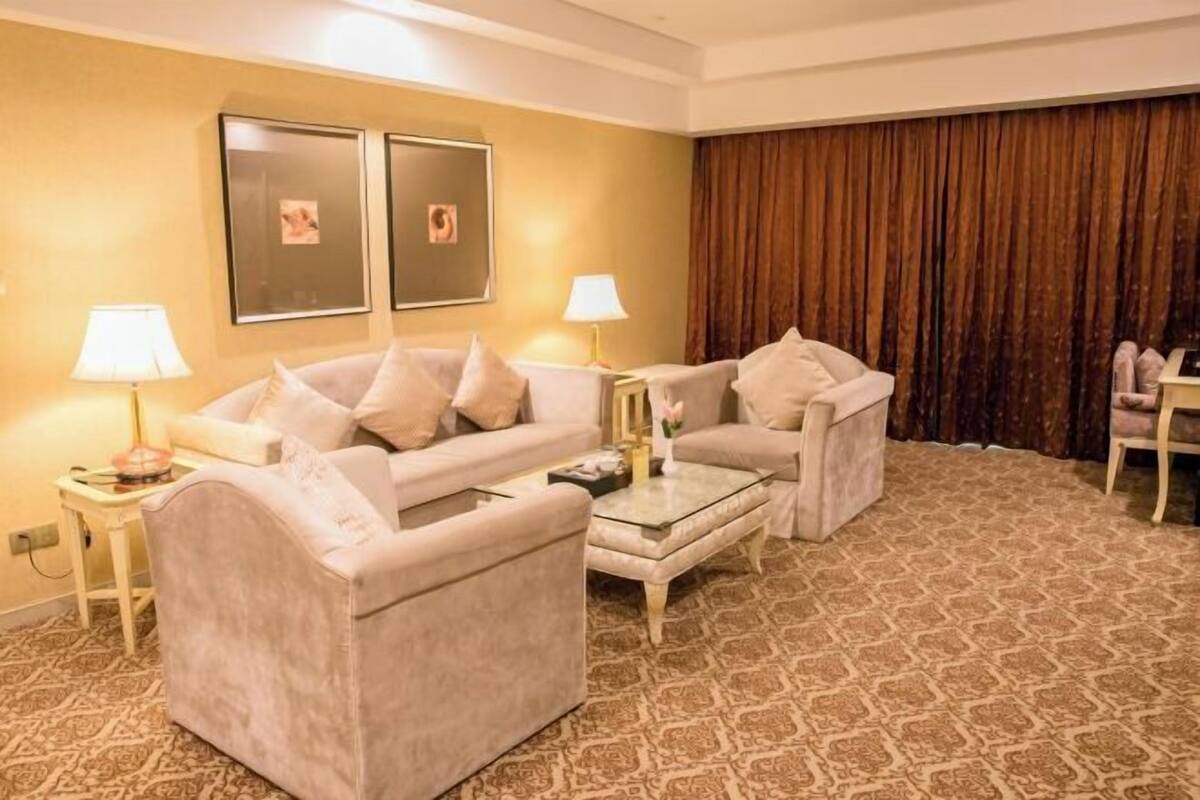 A hotel room with a beige sofa set, a glass coffee table, a patterned carpet, a large window with curtains, and framed pictures on the wall.