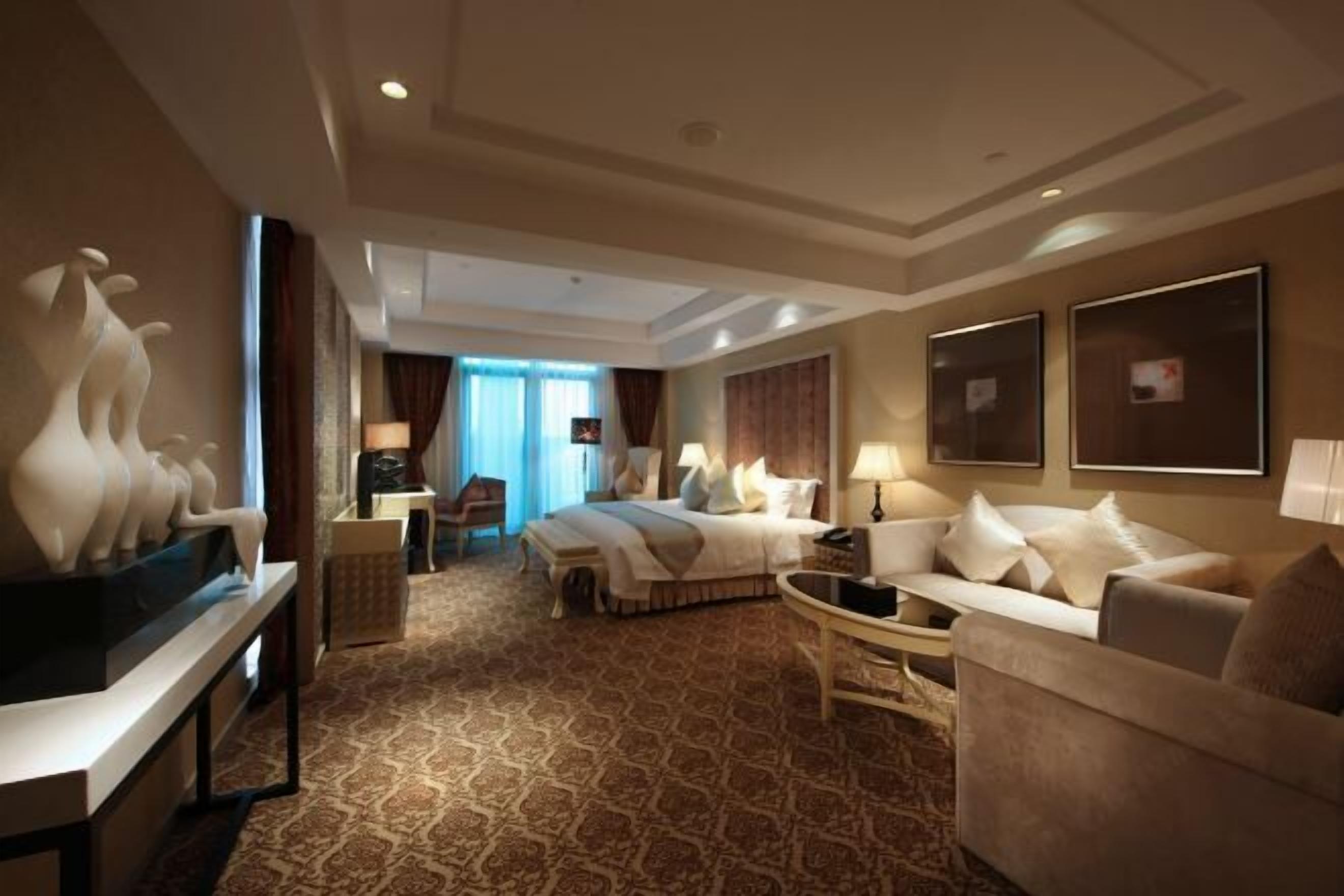 Habitación de hotel con cama, sofá, mesa de centro, televisor y escritorio con lámpara.