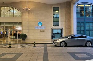 Entrada de un edificio con un coche estacionado adelante, un letrero con un círculo azul y una planta en maceta.