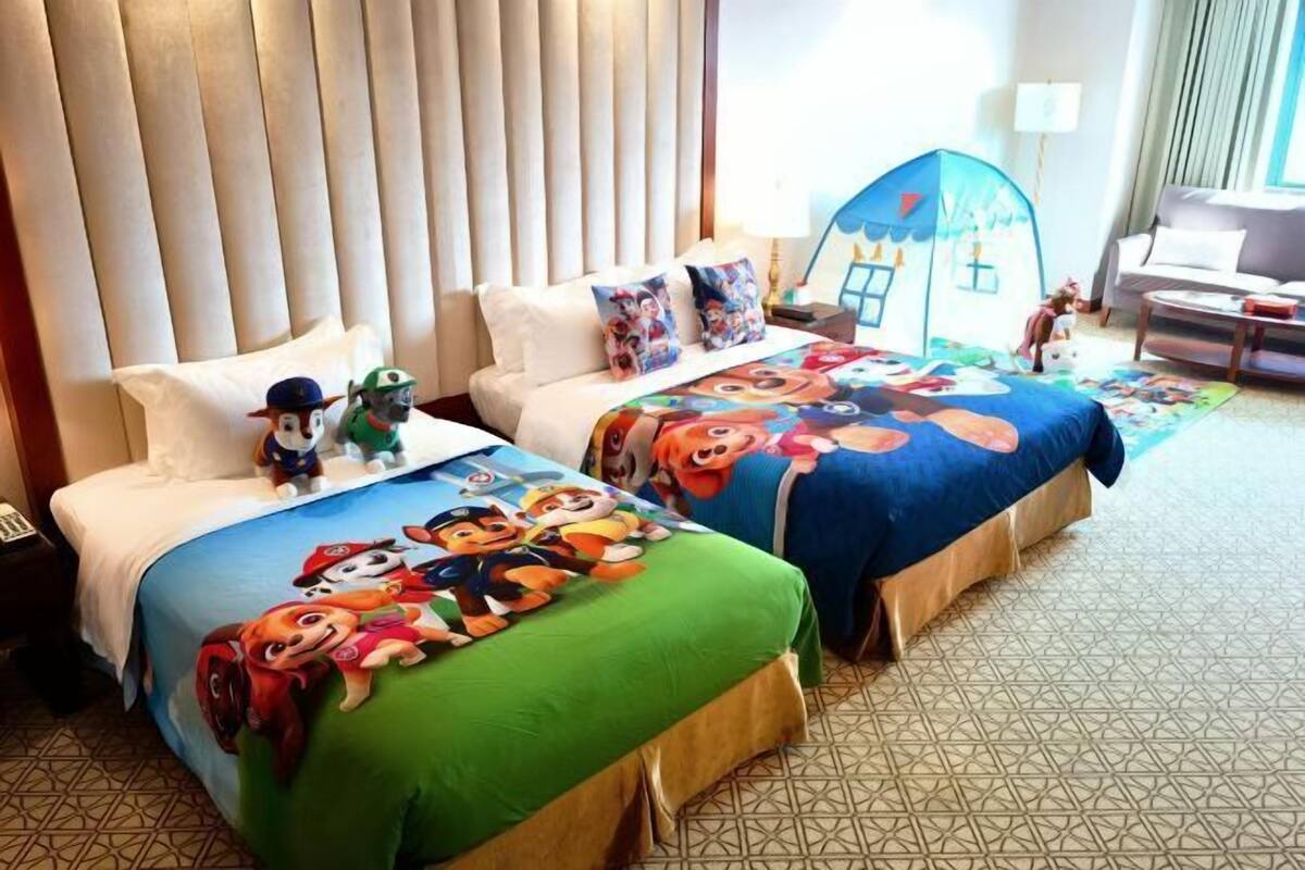 A hotel room with two beds, each with a colorful bedspread featuring cartoon characters. There is a play tent in the room.