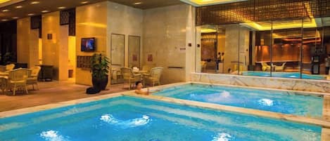 A hotel swimming pool area with multiple pools, lounge chairs, and a television screen.