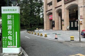 Un panneau vert avec du texte blanc indiquant une nouvelle station de recharge pour véhicules électriques, une entrée de bâtiment et une zone de stationnement délimitée par des barrières rayées de jaune et de noir.