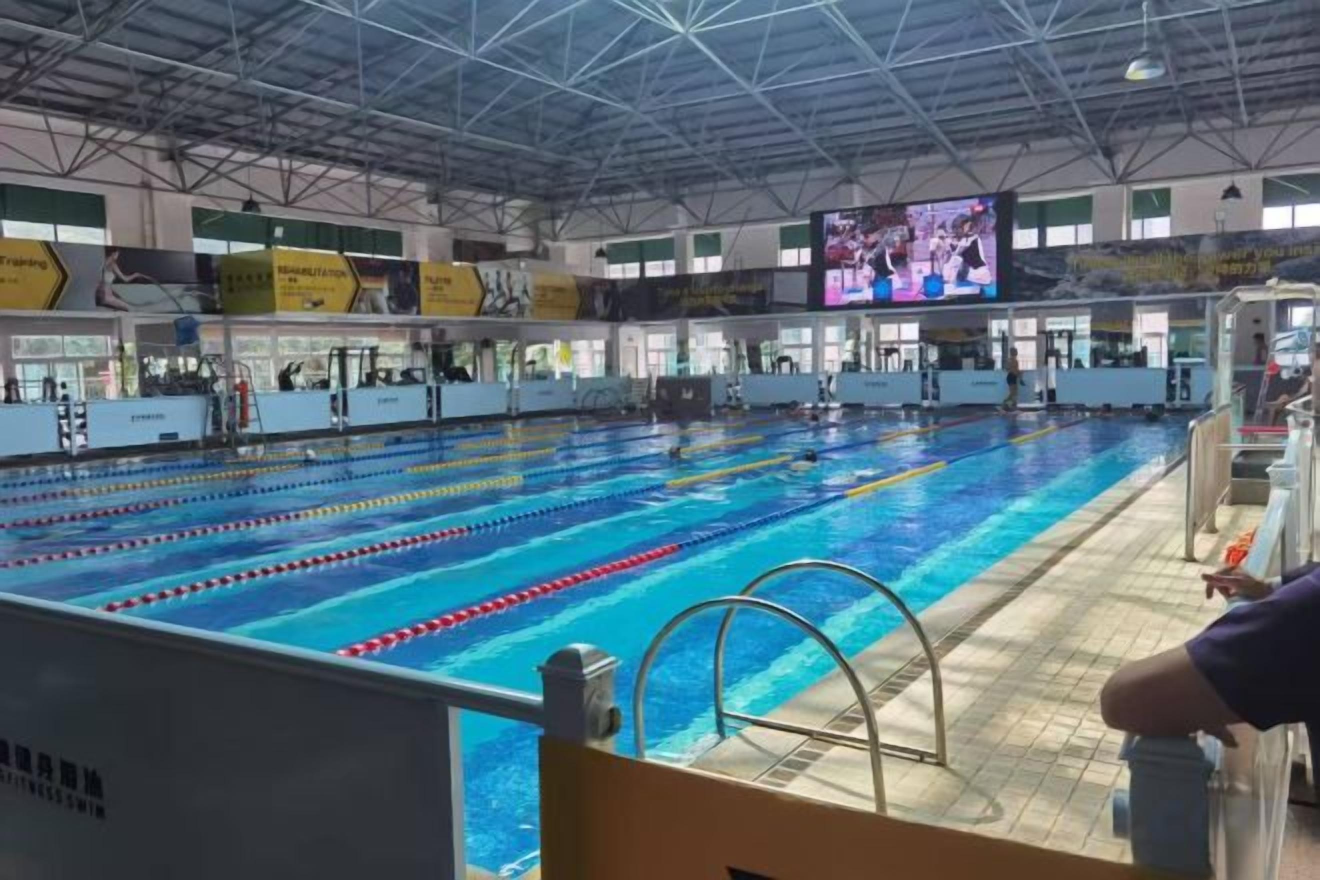 An indoor swimming pool with multiple lanes, a diving platform, and a large screen displaying an event.