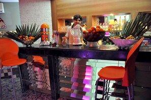 Zona de bar con mostrador, una licuadora y un bol de fruta. Hay sillas anaranjadas y un suelo con estampado de cuadros.