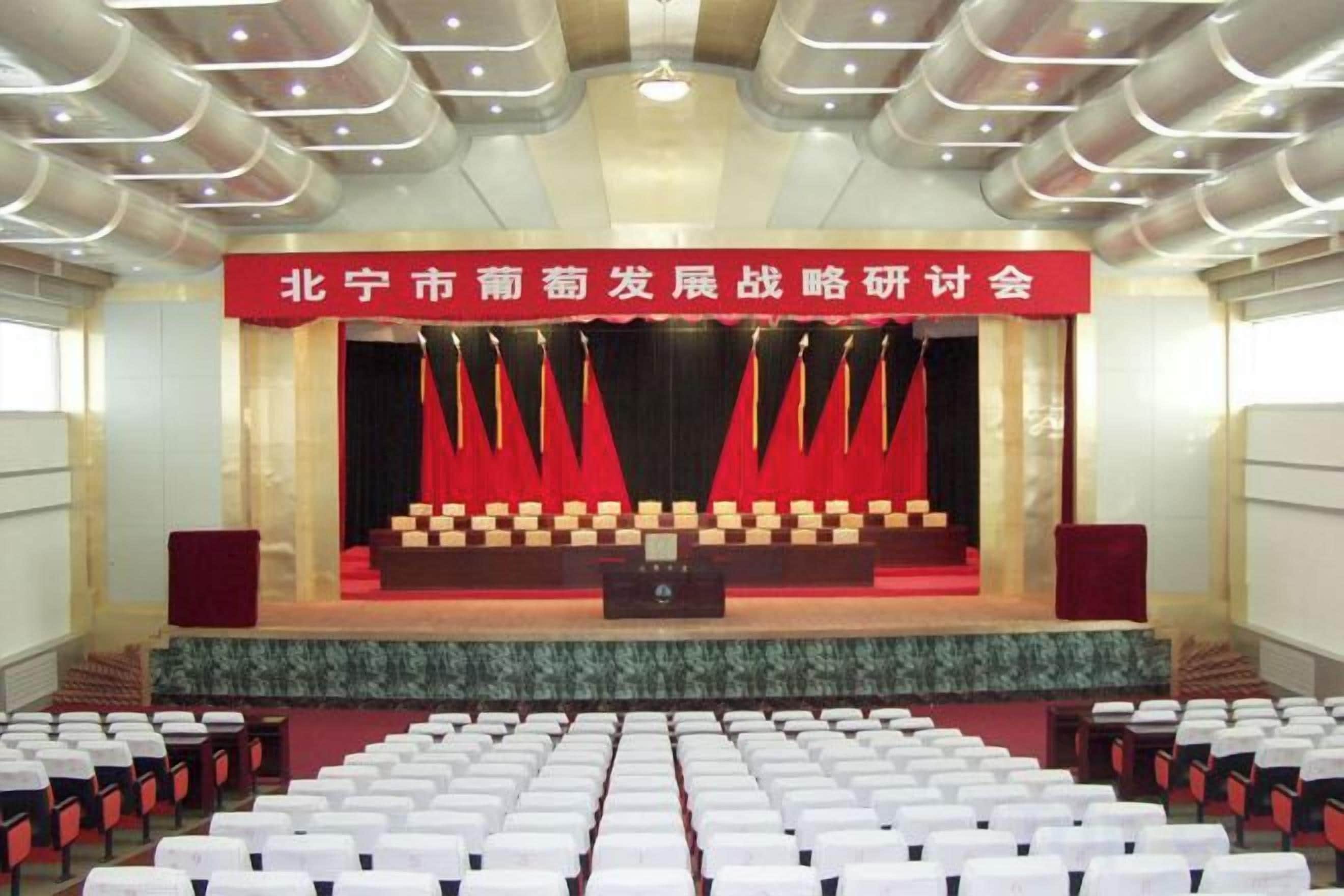 A conference room with rows of white chairs, a stage with a red curtain, and a banner above the stage.