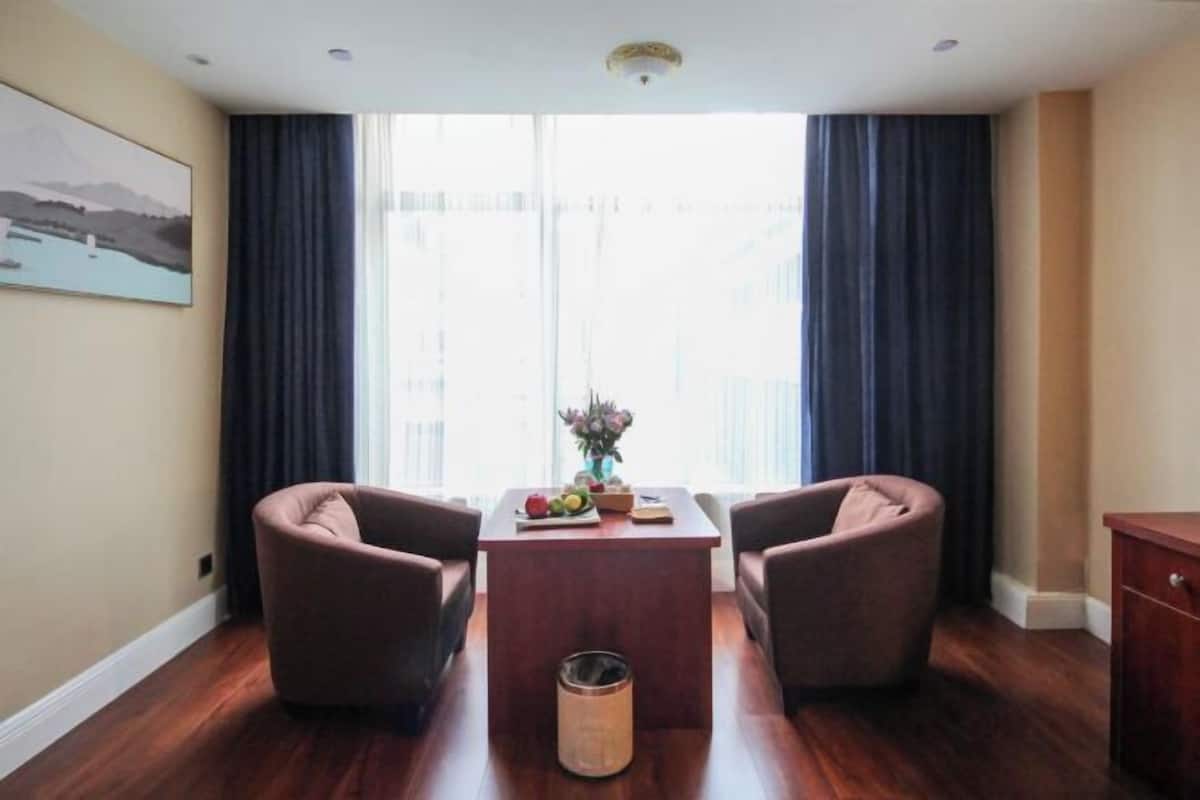 Ein Zimmer mit zwei braunen Sesseln, einem Holztisch mit Obst und einer Vase, sowie einem Fenster mit Vorhängen.