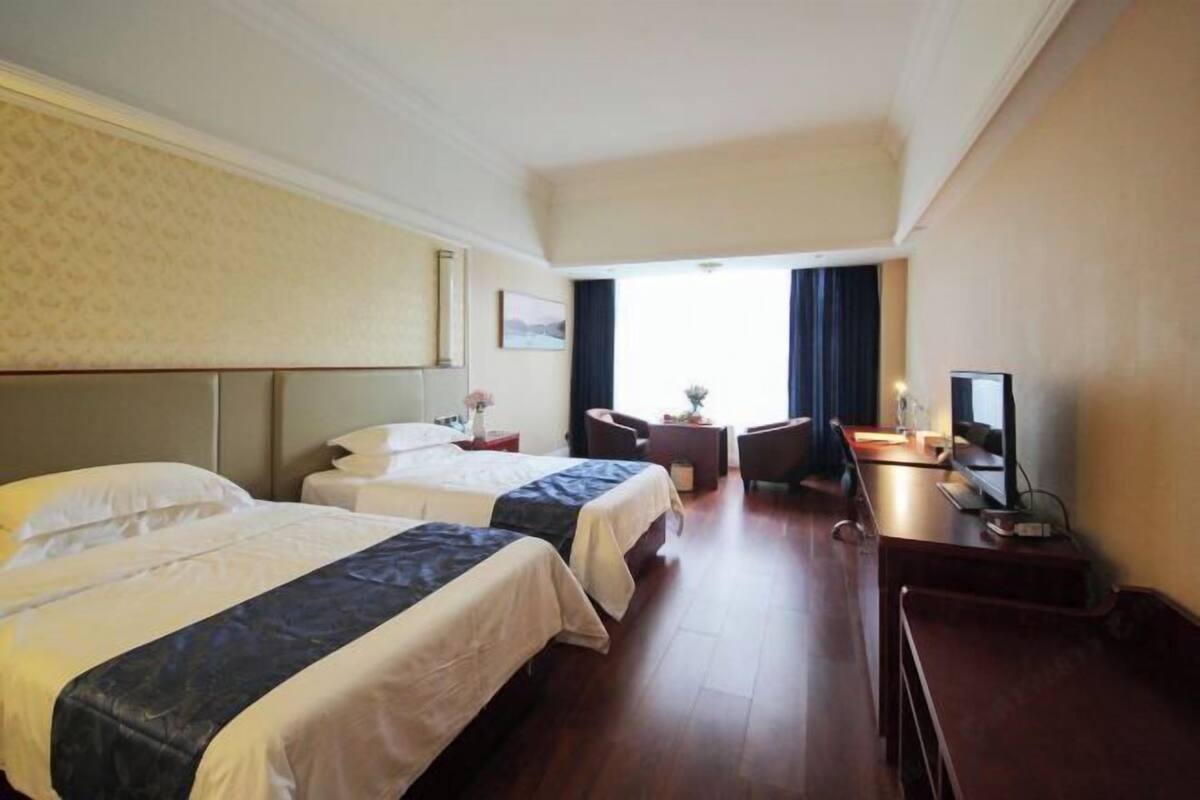 Ein Hotelzimmer mit zwei Betten, einem Schreibtisch, einem Stuhl, einem Fernseher und einem Fenster mit Vorhängen.