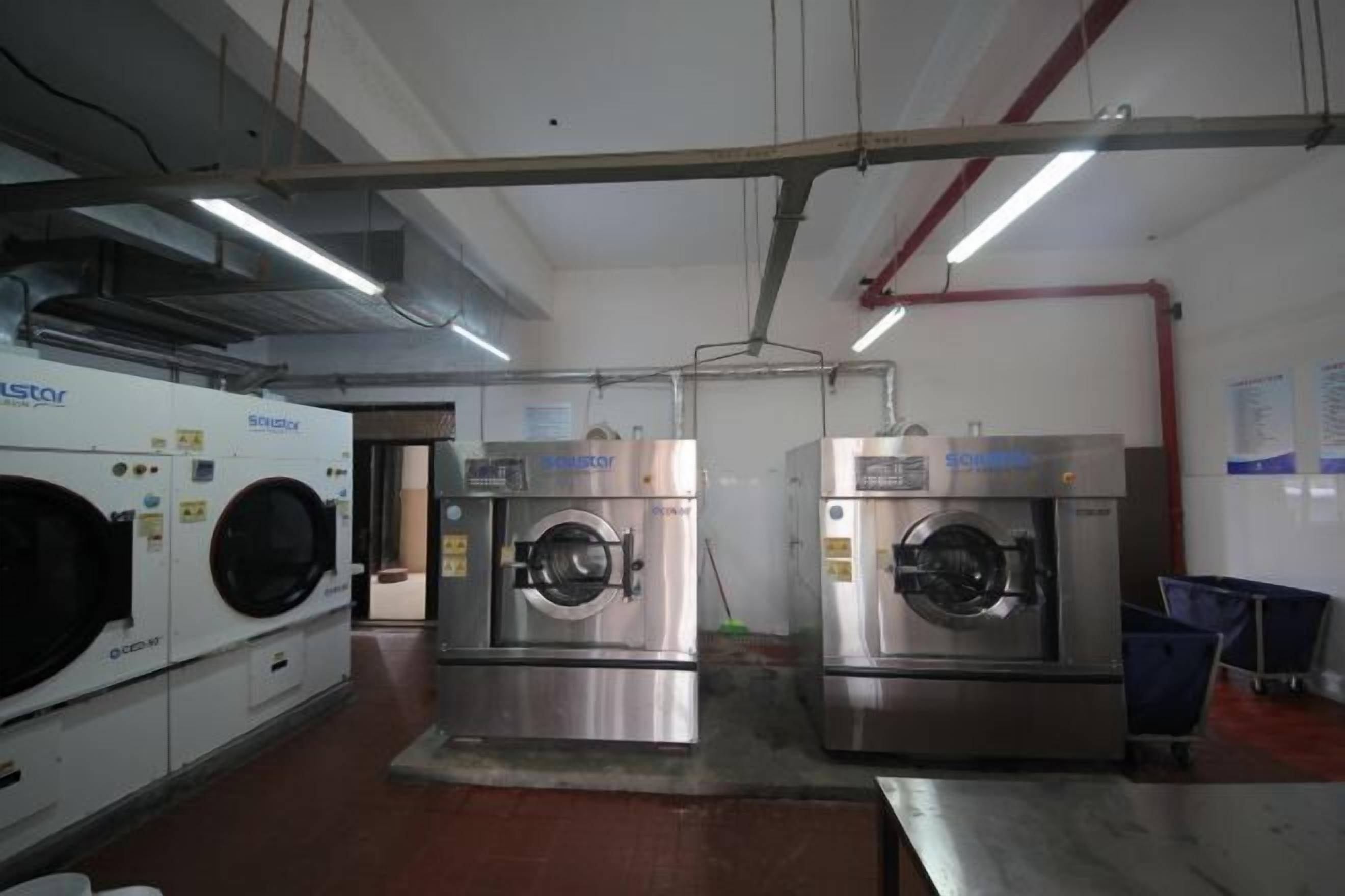 A laundry facility with industrial washing machines and dryers, metal flooring, and visible plumbing and electrical systems.
