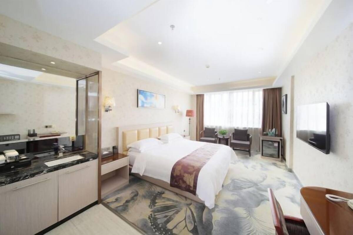 A hotel room with a bed, a desk, a chair, a TV, and a bathroom with a mirror and sink.