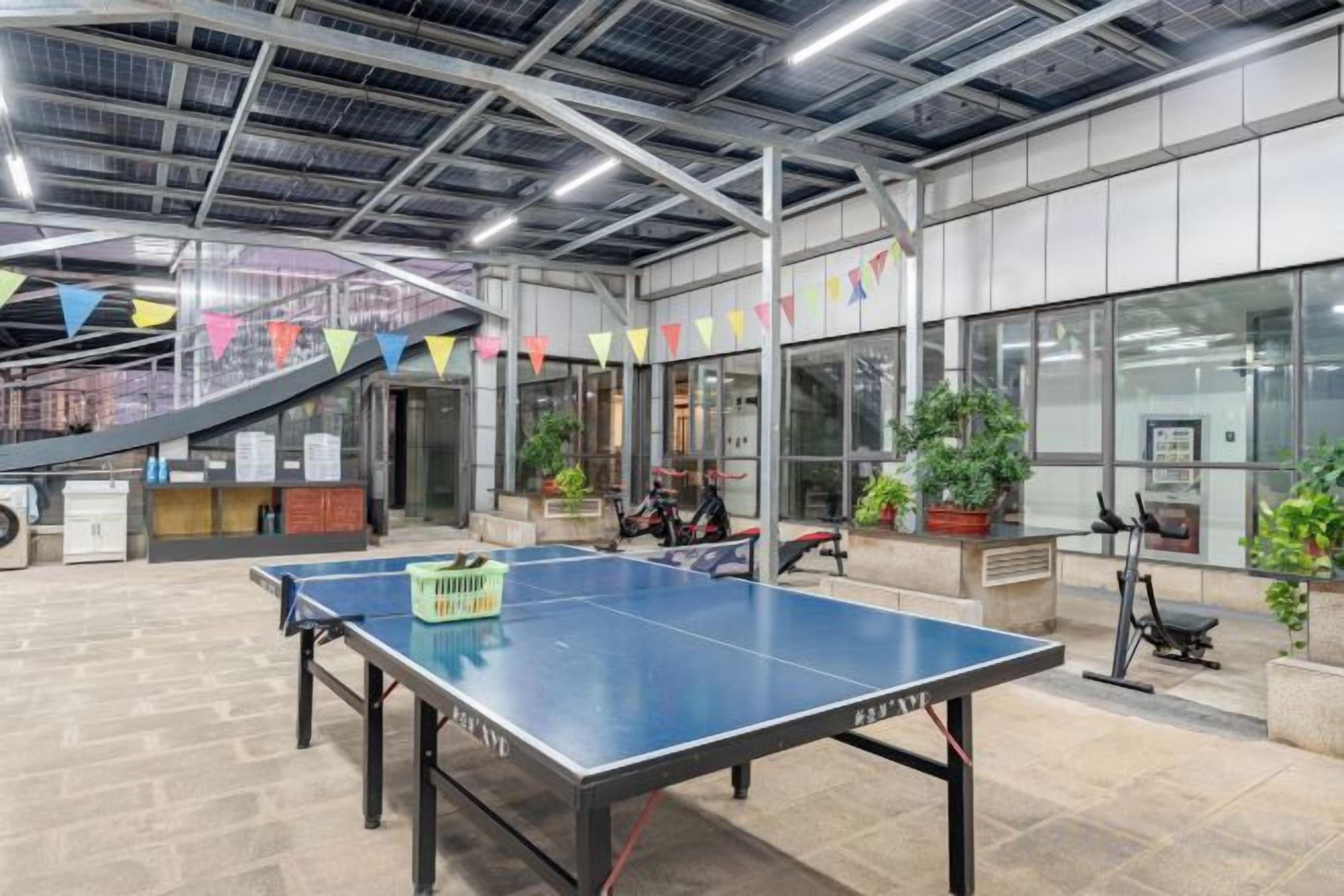 A ping pong table in a spacious indoor area with a staircase, potted plants, and exercise equipment.