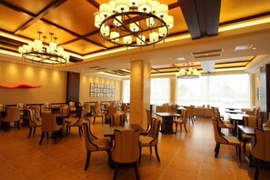 Un comedor amplio en un hotel, con varias mesas y sillas, grandes candelabros y vistas de la ciudad a través de amplias ventanas.