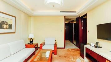 A hotel room with a sofa, armchair, coffee table, TV, and a desk with a flat-screen TV.