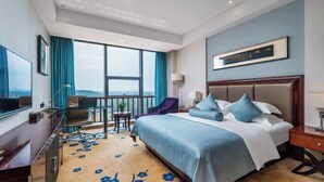 Ein Hotelzimmer mit einem großen Bett, einem Schreibtisch, einem Sessel, einem Fernseher und einem Balkon mit Blick.