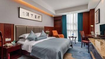 A hotel room with a large bed, a desk, and a chair. There is a nightstand with a phone and a lamp. The room has a carpet and a window with curtains.