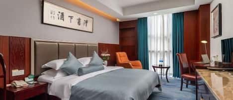 A hotel room with a large bed, a desk, and a chair. There is a nightstand with a phone and a lamp. The room has a carpet and a window with curtains.