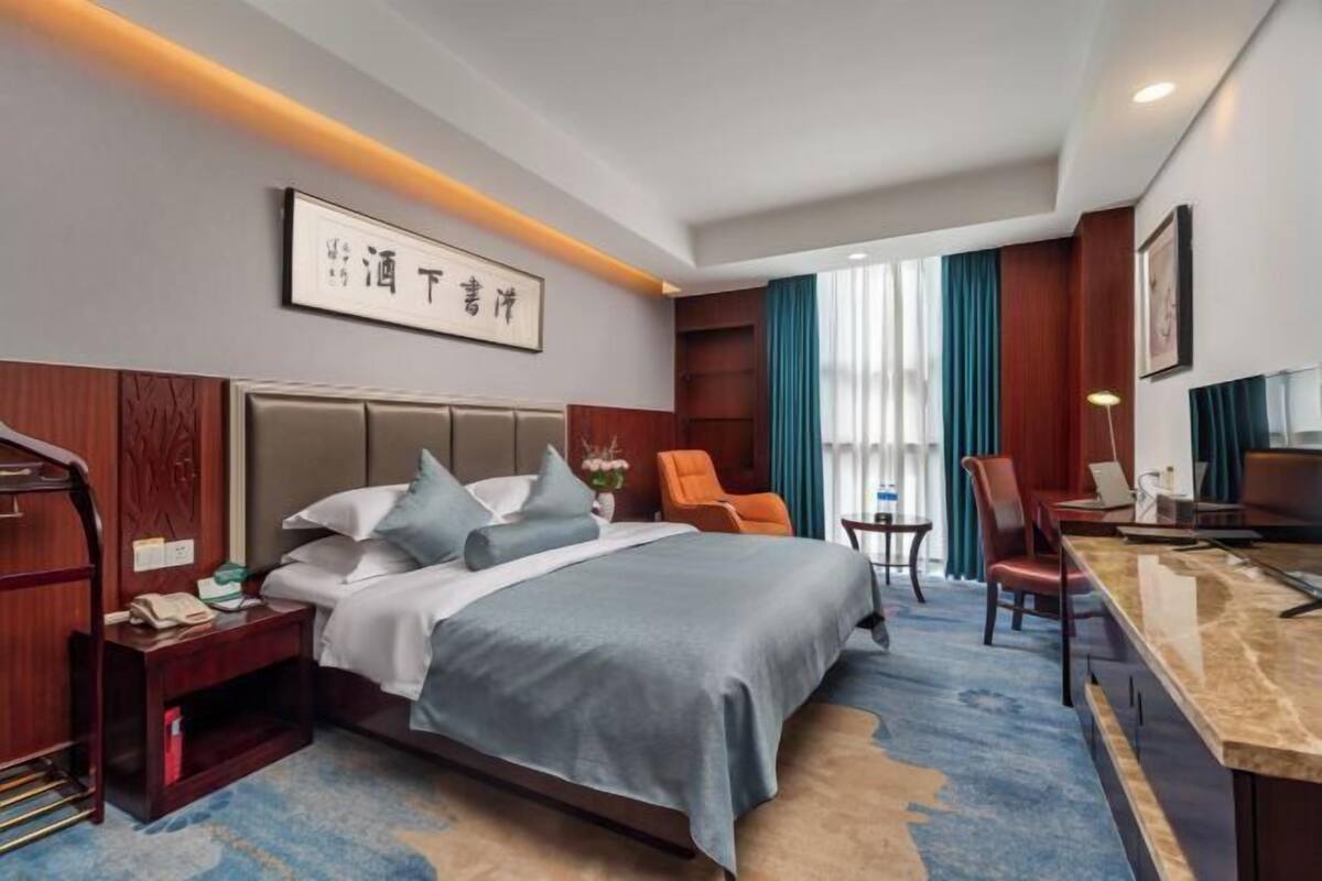 A hotel room with a bed, nightstand, desk, chairs, and a large window with curtains.